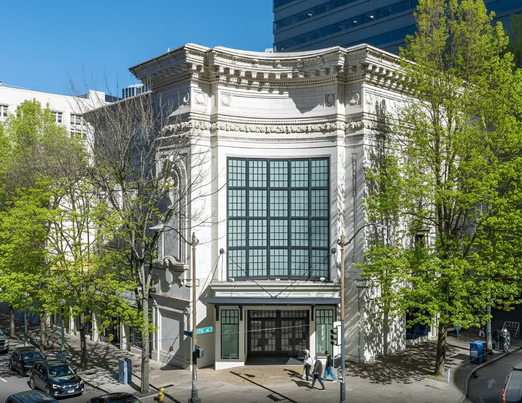 Wide street view of the full historic Coliseum facade framed by green trees and downtown buildings in Seattle