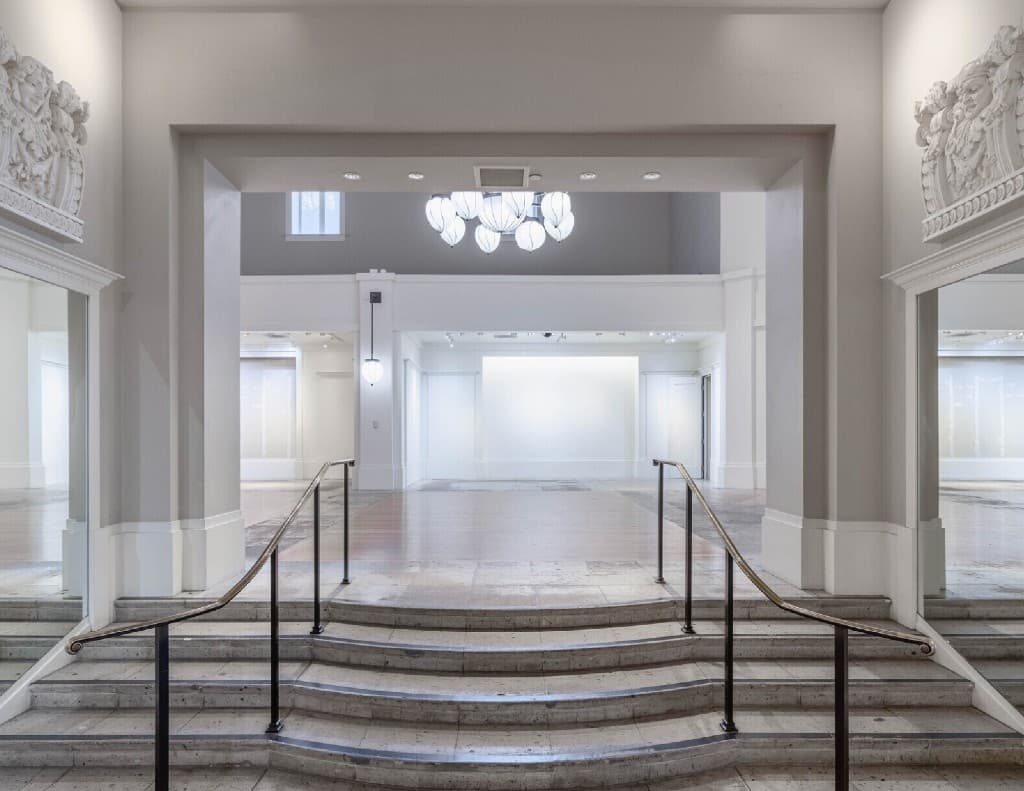 The historic Coliseum interior, looking over curved stone steps and black handrails into a white gallery hall under modern pendant lighting